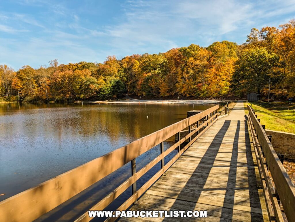 A wooden walkway leading over a lake at Colonel Denning State Park in Cumberland County, PA, with autumn's full display in the background. Trees with leaves in shades of orange, yellow, and green reflect in the still water, under a clear blue sky, creating a peaceful and scenic pathway.