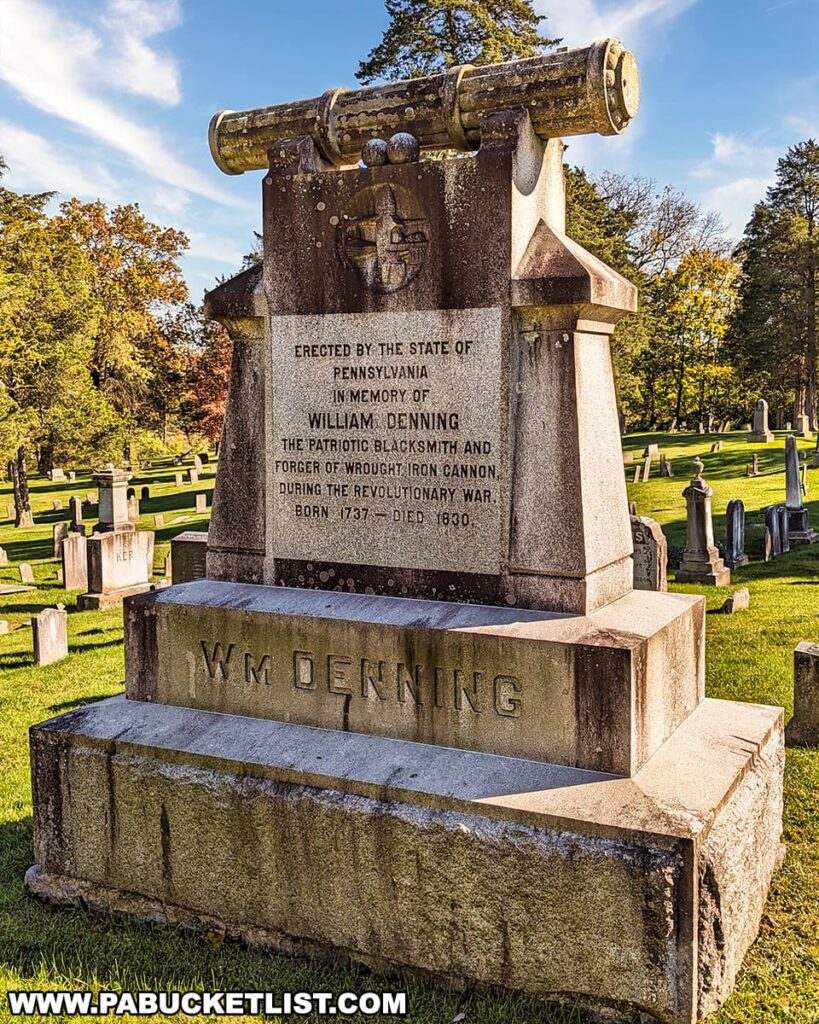 The photo shows the grave of William Denning at Big Spring Presbyterian Church Cemetery in Newville, PA, featuring a distinctive monument erected by the State of Pennsylvania. The monument includes an engraved stone with a wrought-iron cannon atop, commemorating Denning as the patriotic blacksmith and forger of cannons during the Revolutionary War. It includes his birth and death years, 1737 and 1830, respectively. The image captures the monument in sharp detail against a backdrop of a bright blue sky and autumn trees, with the cemetery's peaceful expanse visible in the background.