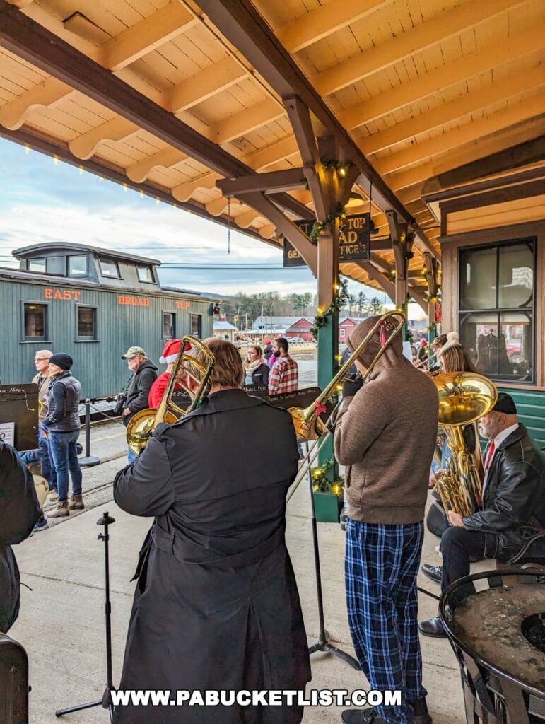 A brass band performs at the East Broad Top Railroad station in Huntingdon County, Pennsylvania. Musicians with trombones and a tuba play under the wooden shelter of the station, adorned with Christmas garland and lights. Passengers and onlookers gather in the background, enjoying the festive music in the outdoor setting.
