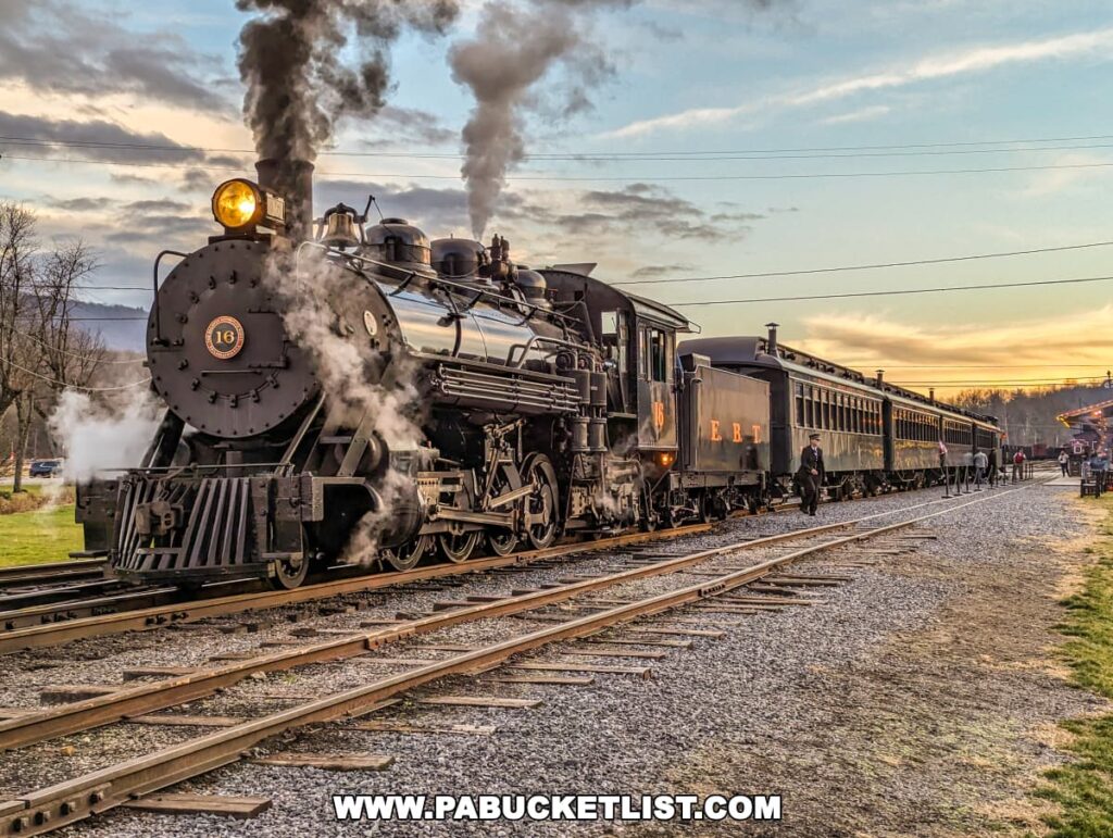 A beautifully preserved steam locomotive, number 16, of the East Broad Top Railroad in Huntingdon County, Pennsylvania, is captured during an evening run. The train, with its headlight aglow, emits billowing clouds of steam against the backdrop of a sunset sky. Period-clad railway staff are seen on the ground, contributing to the historical ambiance of the scene.