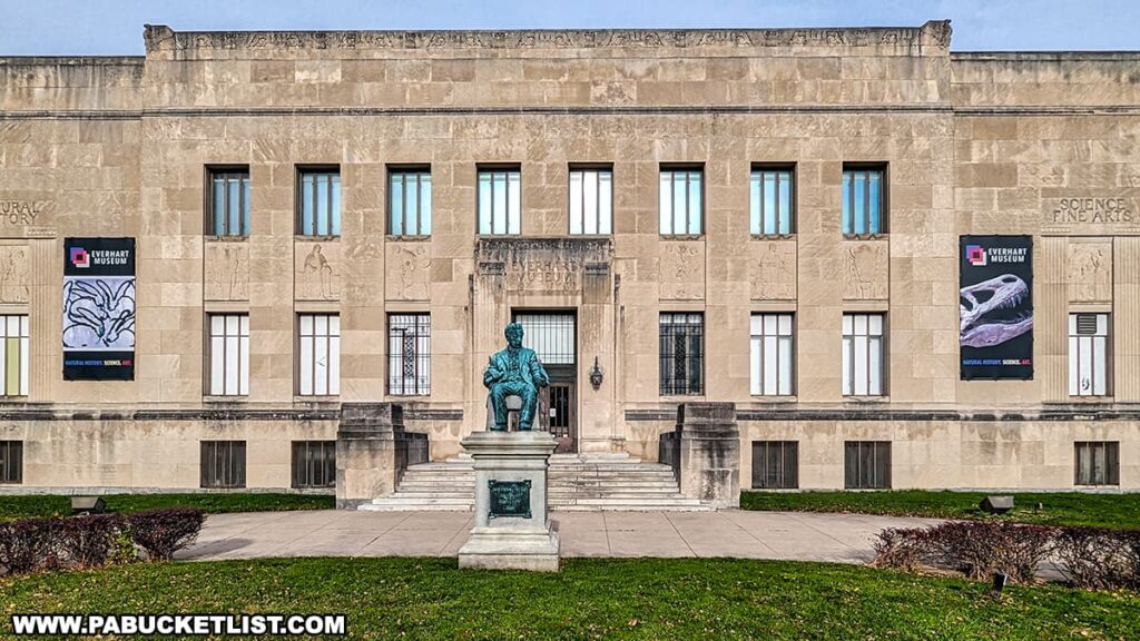 The front entrance of the Everhart Museum located in Nay Aug Park, Scranton, Pennsylvania. The museum's facade is adorned with decorative carvings and two large banners promoting natural history and art exhibitions. In front of the entrance stands a bronze statue on a pedestal, with neatly maintained grass and clear skies in the background.