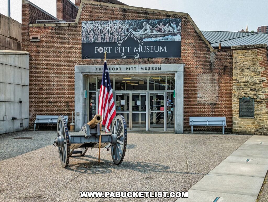The entrance of the Fort Pitt Museum in Pittsburgh, Pennsylvania, with an antique cannon in the foreground displaying the American flag. The museum's facade features a brick wall and a banner with an image of colonial figures, welcoming visitors to explore the historical site.