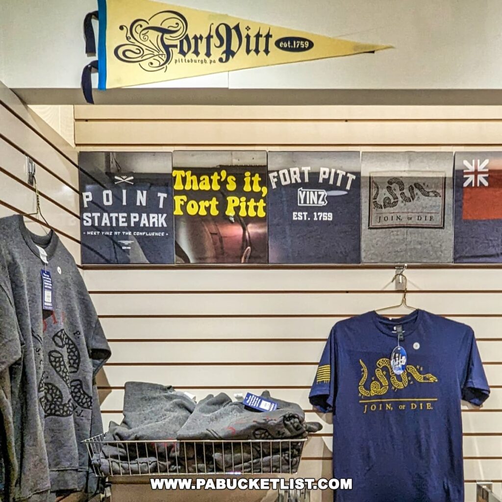 Merchandise display at the Fort Pitt Museum gift shop in Pittsburgh, Pennsylvania, featuring a variety of themed t-shirts with slogans and images related to Fort Pitt and Point State Park, as well as a yellow pennant with 'Fort Pitt est. 1759' written on it.