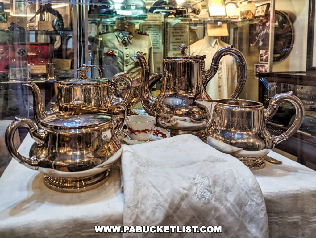 A close-up view of a gleaming vintage silver tea service set on display at the Gettysburg Museum of History. The collection includes a teapot, coffee pot, creamer, and sugar bowl, all reflecting the lights of the museum. In the background, through the glass of the display case, other historical memorabilia can be partially seen. A white linen cloth with an embroidered emblem is draped in the foreground.