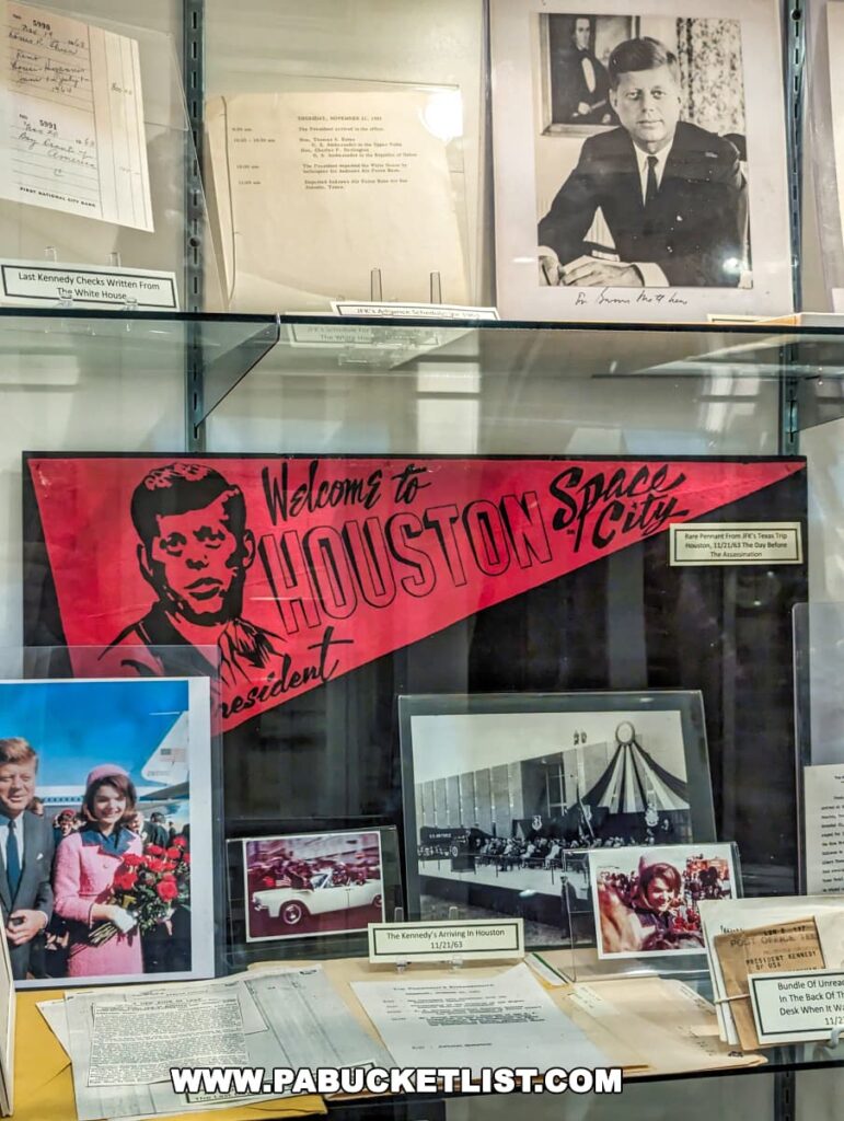 A collection of John F. Kennedy memorabilia displayed at the Gettysburg Museum of History. Items include a large banner with the words 'Welcome to Houston Space City' featuring an illustration of JFK, alongside photographs of Kennedy, including one with a signature. Documents below the photos include a note identified as the 'Last Kennedy Check Written From The White House' and other papers relating to JFK. In the bottom right, a caption states 'The Kennedy's Arriving in Houston 11/21/63'.