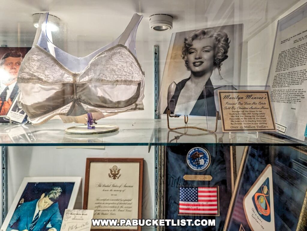 Exhibit at the Gettysburg Museum of History featuring Marilyn Monroe memorabilia. A large brassiere is centrally displayed, with an accompanying placard stating it's from the estate sale by Christie's auction house. To the right is a framed photograph of Monroe with a radiant smile. Below the display case, other historical items are visible, including a framed presidential seal.