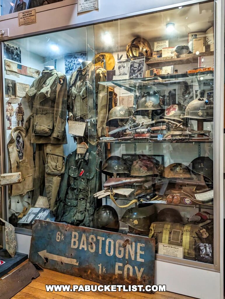 A comprehensive military memorabilia display at the Gettysburg Museum of History. The collection includes World War II military uniforms, helmets, and equipment, with a sign indicating 'Bastogne 1k FOY'. Each artifact appears to have a descriptive note, providing context and history. In the foreground, the blue road sign 'Bastogne 6k FOY' adds to the authenticity of the exhibit.