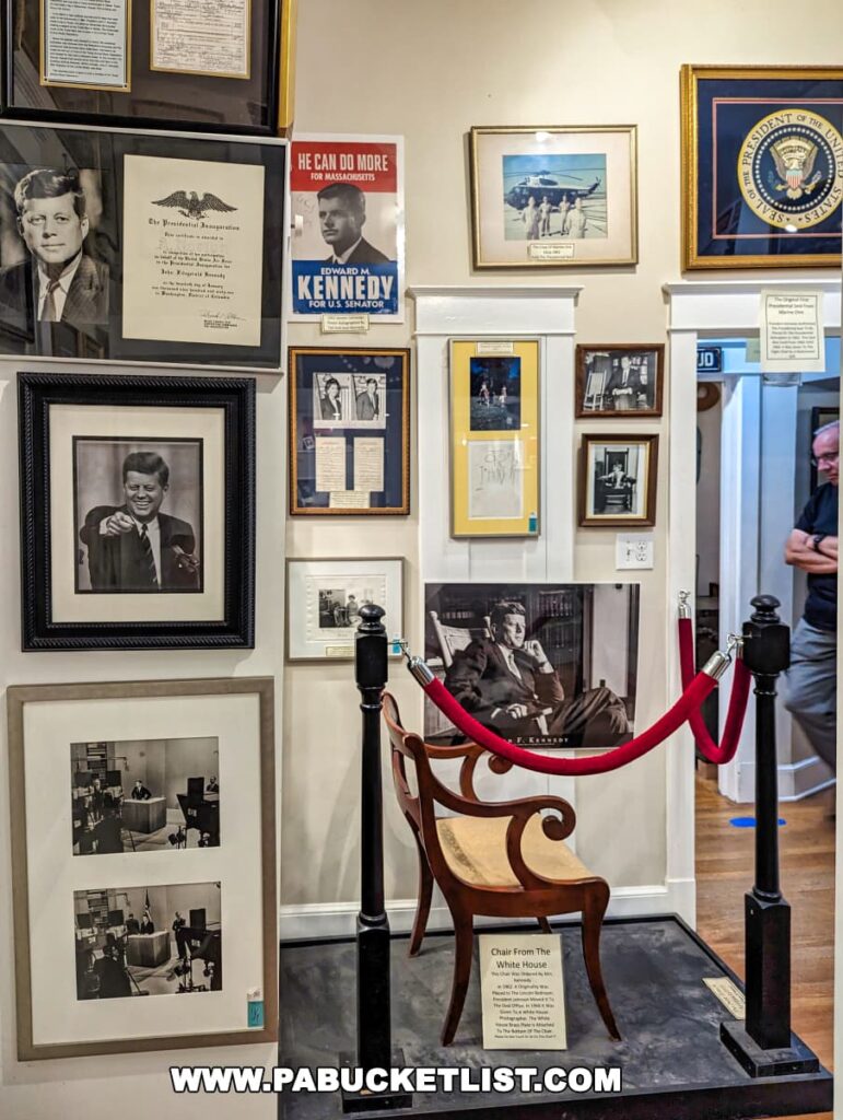An exhibit dedicated to President Kennedy at the Gettysburg Museum of History. The display includes framed photographs, campaign posters, and historical documents on the walls, and a chair from the White House cordoned off with a red velvet rope. One notable poster reads 'He Can Do More For Massachusetts' for Edward M. Kennedy for U.S. Senator. A presidential seal hangs on the upper right.