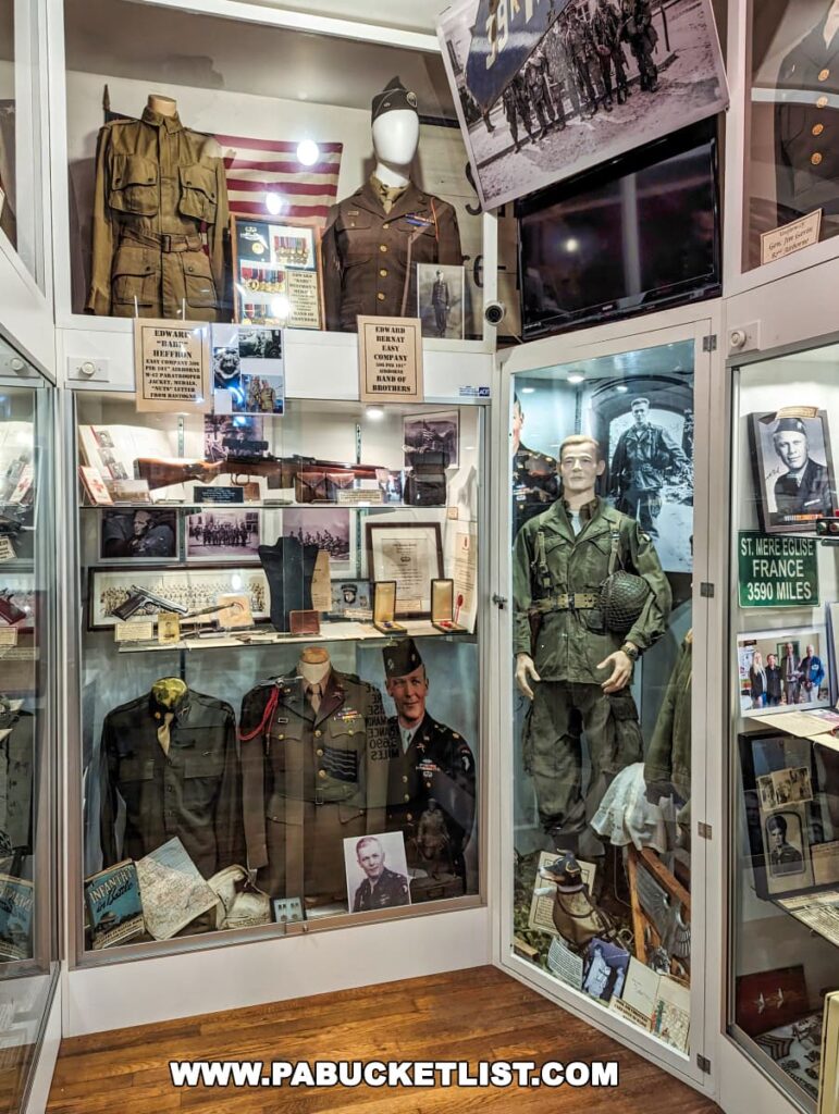 A display of World War II military uniforms and memorabilia at the Gettysburg Museum of History. Several mannequins are dressed in period uniforms, complete with insignia, medals, and headgear. Accompanying the uniforms are photographs, documents, and personal items from soldiers, with descriptive notes providing context. A sign indicating 'E Company 506th PIR' references the 'Band of Brothers.' The exhibit is a tribute to the individuals and units that served.