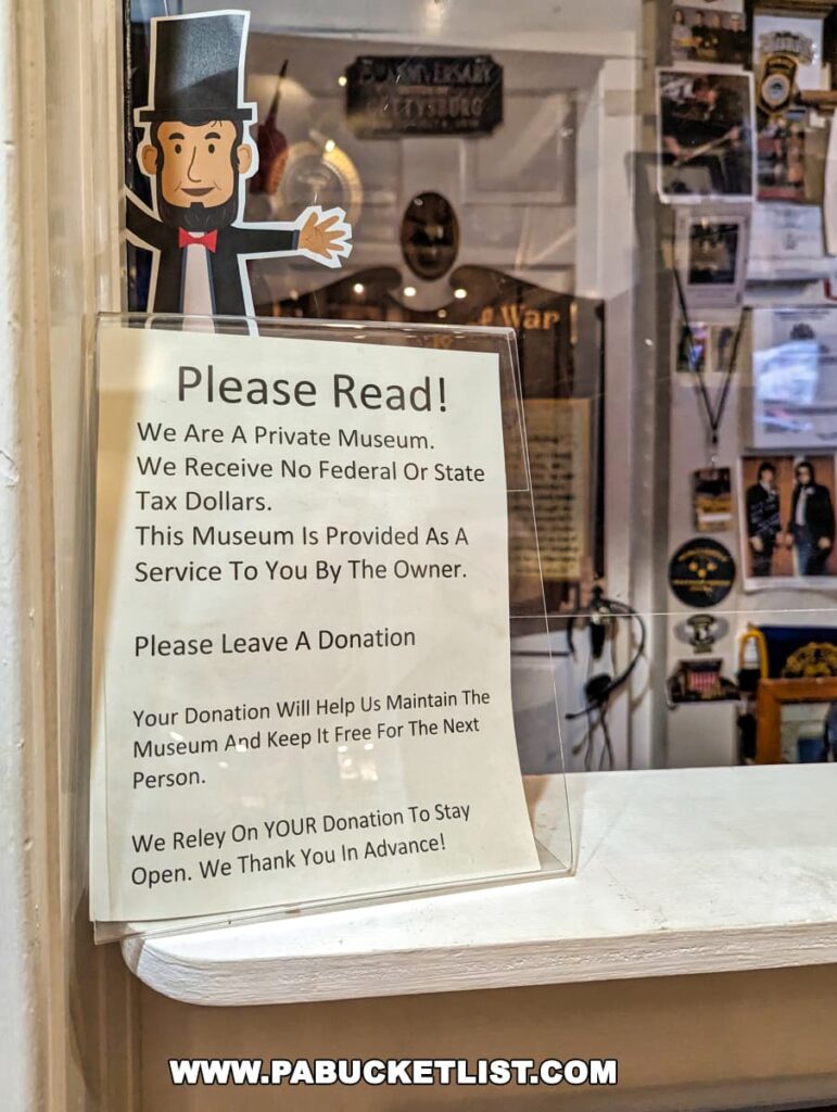 Signage at the Gettysburg Museum of History requesting donations. The notice, placed on a window ledge, features a graphic of Abraham Lincoln with an outstretched hand, accompanied by text appealing to visitors for support, stating the museum is privately funded and relies on donations to remain open and free for others. In the background, glimpses of Civil War exhibits and memorabilia are visible.