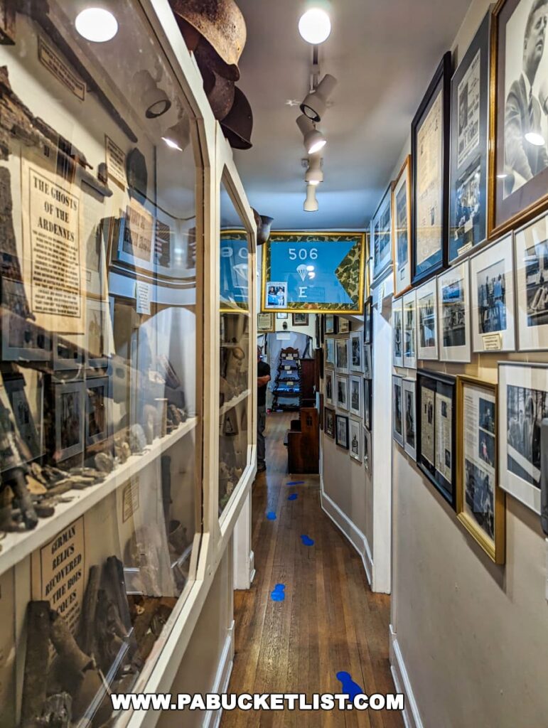 A narrow corridor inside the Gettysburg Museum of History lined with floor-to-ceiling exhibits. The left wall features a glass case filled with historical artifacts, above which hang helmets and descriptive texts. The right wall is adorned with framed photographs and documents, including a prominent blue banner for the '506th V E' with a background of clouds and sky. Directional blue footprints are placed on the wooden floor, guiding visitors through the museum. Overhead lighting illuminates the space.