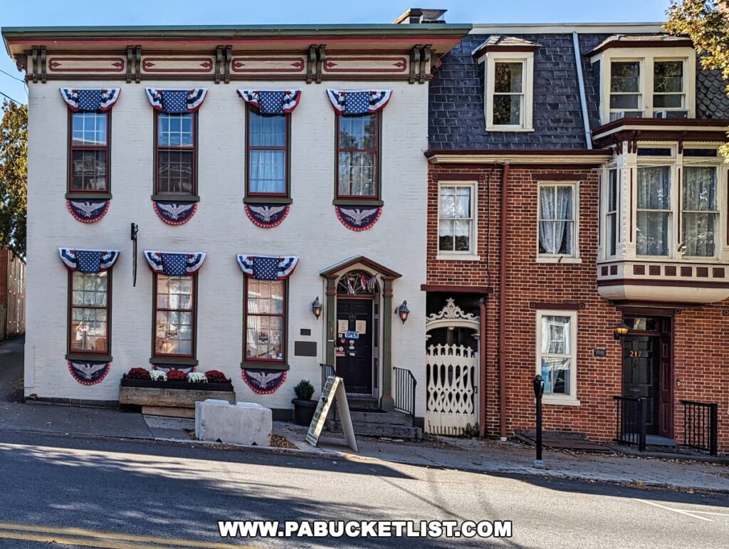 The front façade of the Gettysburg Museum of History in Gettysburg, Pennsylvania. The two-story white building is decorated with patriotic bunting on its windows, reflecting its historical significance. Adjacent is a red brick building with traditional architecture, featuring a bay window and a white decorative fence. A sign indicating the museum is open hangs beside the entrance door, and a sidewalk leads up to the buildings.