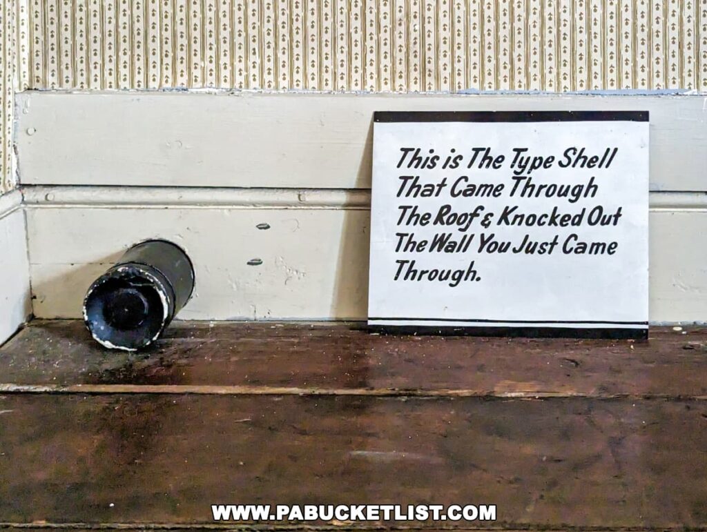 A photo from the Jennie Wade House in Gettysburg, Pennsylvania, displaying a black Civil War-era shell lying on a wooden floor against a wall with patterned wallpaper. Beside it, a sign explains that this is the type of shell that came through the roof and knocked out the wall visitors have just passed through.