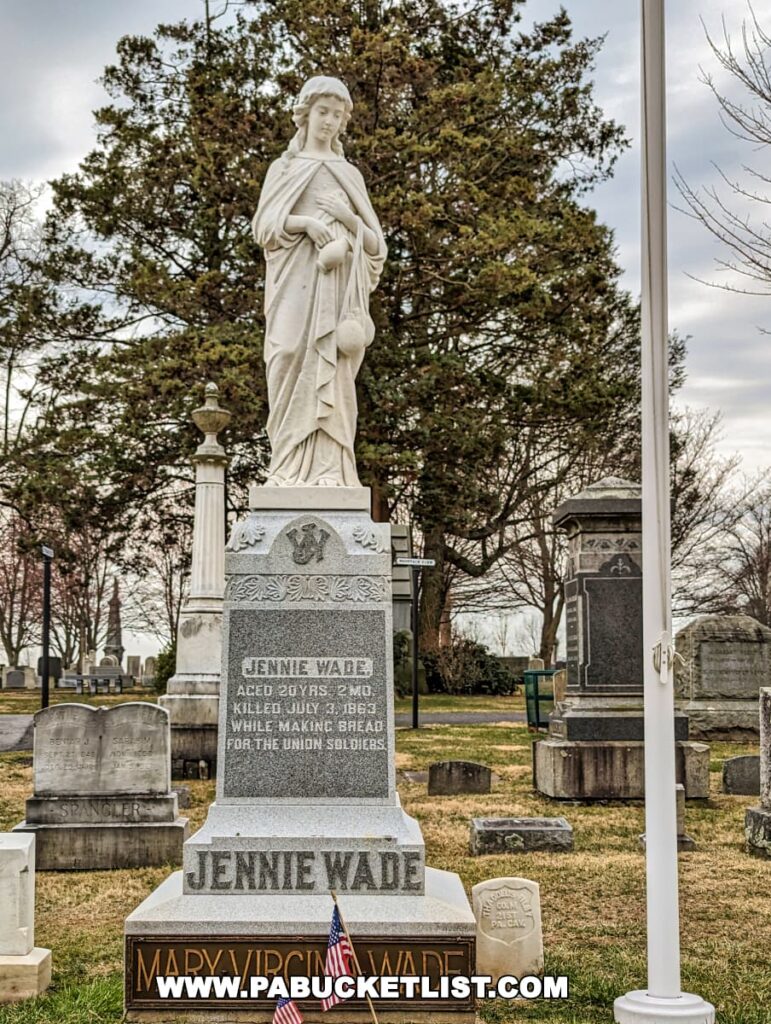 The burial site of Jennie Wade at Evergreen Cemetery in Gettysburg, Pennsylvania, marked by a statue of a woman and a gravestone that reads her name, age, and the fact that she was killed while making bread for Union soldiers. American flags are placed at the base of the monument, with mature trees in the background.