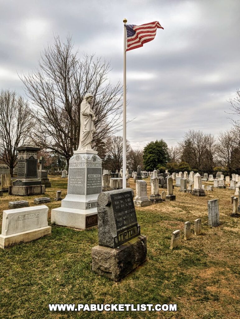 Jennie Wade's gravesite at Evergreen Cemetery in Gettysburg, Pennsylvania, featuring a white monument with a statue on top, and her name inscribed on the base. An American flag flies on a pole in the background, with the cemetery's landscape of headstones stretching out under an overcast sky.