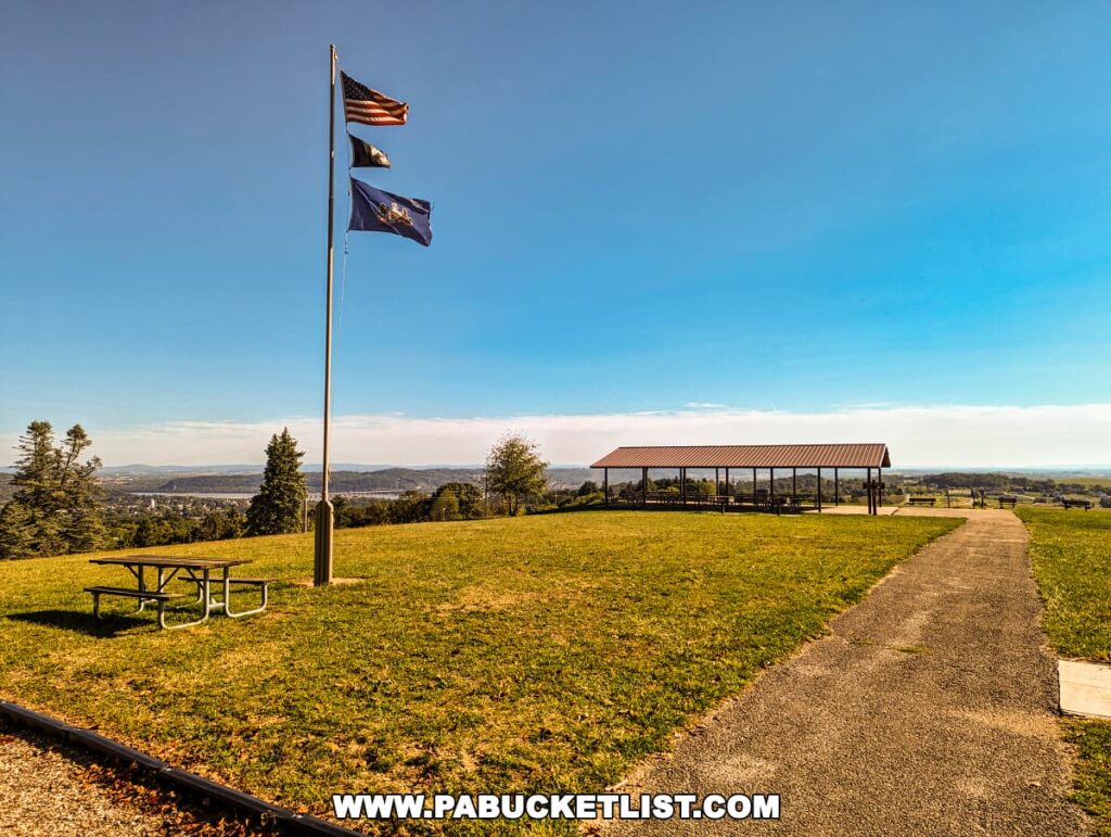 A serene setting at Mount Pisgah Overlook in York County, Pennsylvania, displaying an American flag and a Pennsylvania state flag fluttering in the breeze atop a flagpole. In the midground, a green lawn hosts a picnic table, with a paved pathway leading to a covered pavilion that offers shelter and a place to rest. The landscape stretches into the distance with lush trees and a clear blue sky above.