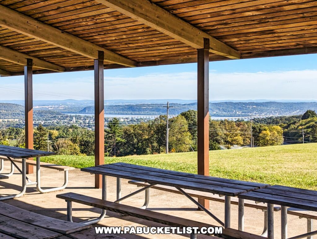 View from a hilltop picnic pavilion at Mount Pisgah Overlook in York County, Pennsylvania. Wooden picnic tables rest under the shelter of the pavilion, with its open sides framing a picturesque view of the rolling hills, trees, and a river in the distance. The bright blue sky and lush greenery evoke a sense of tranquility.