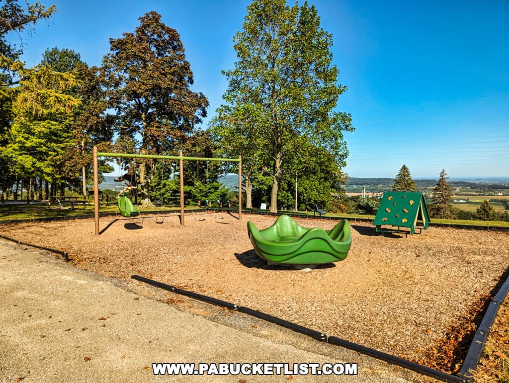 A children's playground at Mount Pisgah Overlook in York County, Pennsylvania, featuring green play equipment on a bed of wood chips. There's a swing set, a wavy slide, and a climbing structure with footholds, set against a backdrop of mature trees and a sweeping view of the valley below. The playground offers a combination of fun and scenic beauty.