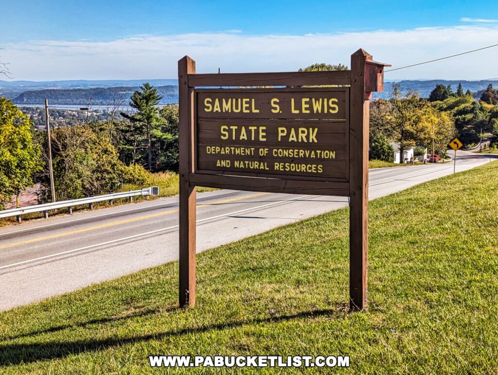 The entrance sign for Samuel S. Lewis State Park in York County, Pennsylvania, set against a clear blue sky. The wooden sign, with carved and painted lettering, indicates the park's affiliation with the Department of Conservation and Natural Resources. Behind the sign, the rolling hills and lush greenery of the area are visible, alongside a road that stretches into the picturesque landscape.
