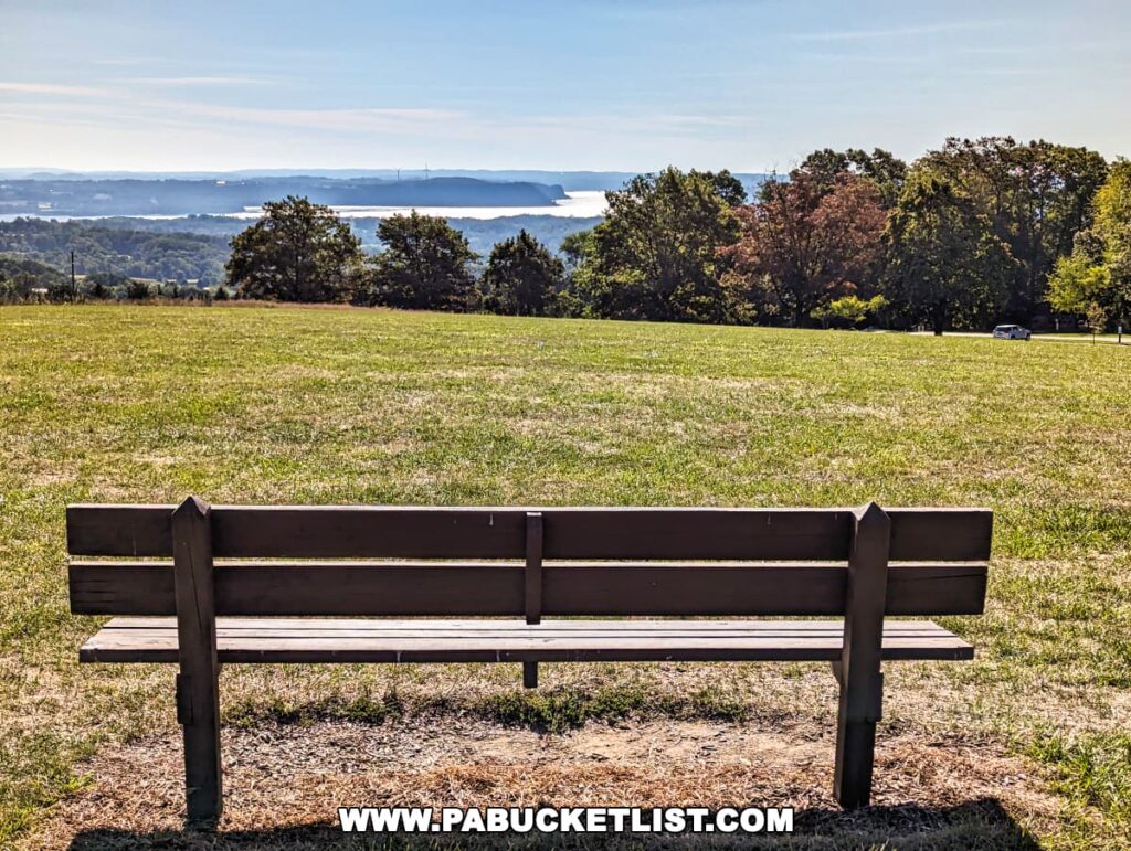 A peaceful scene at Mount Pisgah Overlook in York County, Pennsylvania, showing a solitary wooden bench with a view towards the south over the Susquehanna River. The bench is positioned on a grassy hilltop, inviting visitors to sit and enjoy the expansive view of the river, distant hills, and a clear blue sky. Lush trees frame the edges of the image,