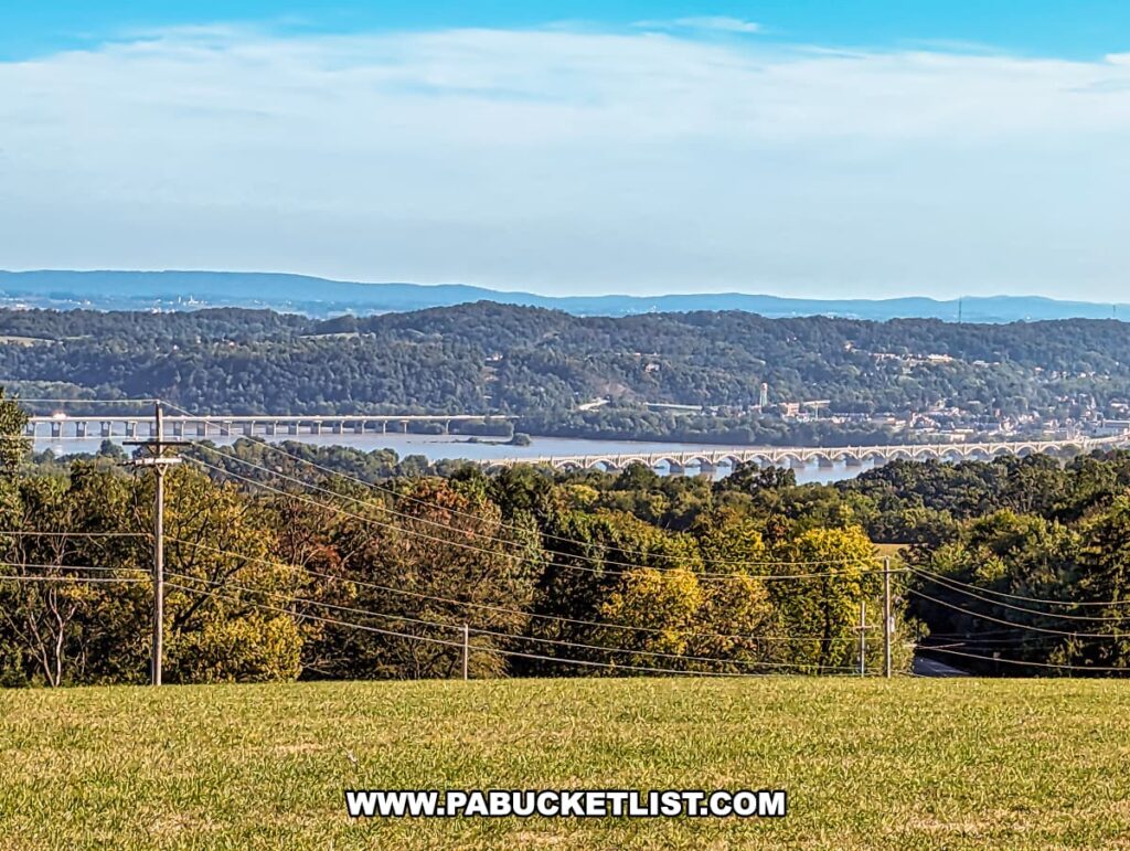 A stunning view from Mount Pisgah Overlook in York County, Pennsylvania, showcasing the Wrightsville-Columbia bridge spanning across the Susquehanna River. The bridge is captured from a distance, with a foreground of a lush grassy field and a backdrop of rolling hills. Power lines slightly intersect the view, leading the eye across the varied greenery and towards the distant waterway.
