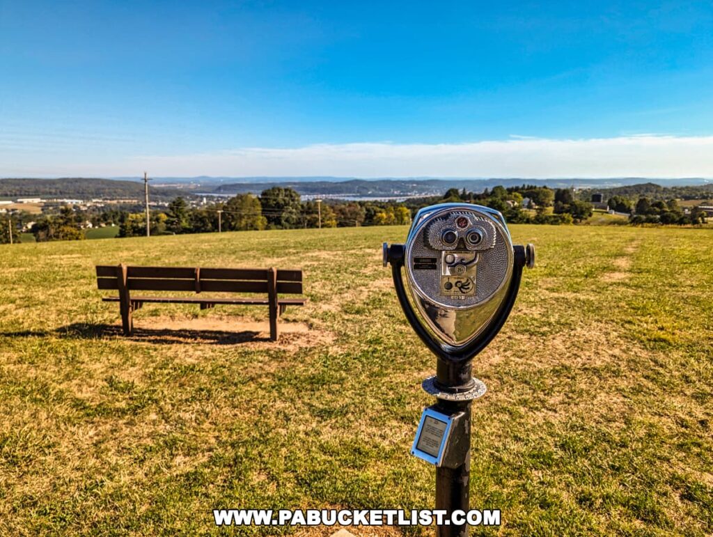 A scenic view from Mount Pisgah Overlook in York County, Pennsylvania, featuring a solitary brown bench facing a vast landscape under a clear blue sky. In the foreground, a coin-operated binocular stand invites visitors to take a closer look at the distant hills and patchwork of fields.