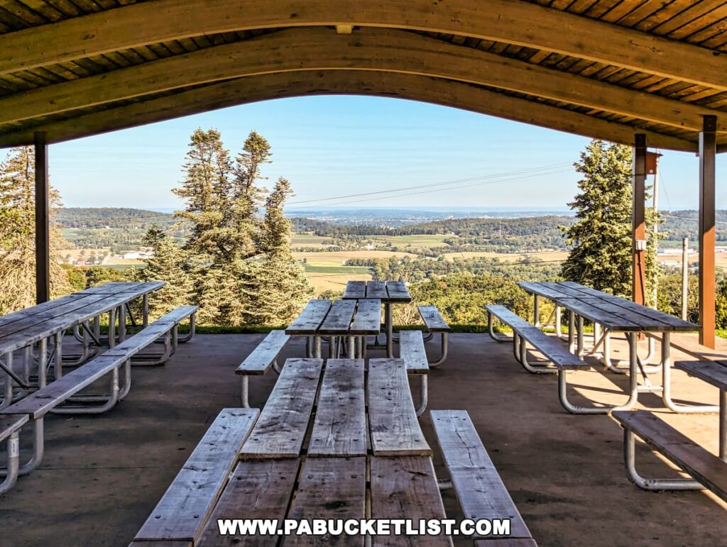 An inviting view from under a curved wooden pavilion at Mount Pisgah Overlook in York County, Pennsylvania, with several picnic tables lined up, ready for visitors. Through the pavilion's open front, one can see a sweeping vista of farmland, patches of forest, and a clear sky. The pavilion serves as a serene spot to enjoy a meal with a view.