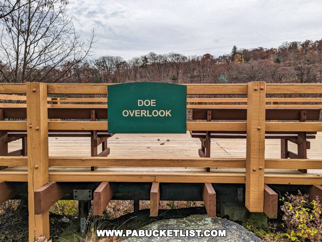 A wooden observation deck at Nay Aug Park in Scranton, Pennsylvania, named 'Doe Overlook.' The deck features sturdy wooden benches and a protective railing, with a green sign indicating the name of the overlook. Leafless trees and a cloudy sky suggest a late fall or early winter season, with a view of the hilly landscape in the background.