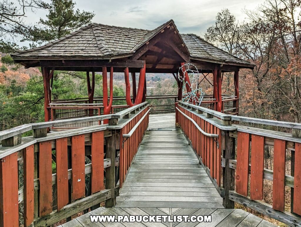 The walkway leading up to the treehouse in Nay Aug Park, Scranton, Pennsylvania, featuring red wooden railings and a weathered wooden floor. The roofed sections of the treehouse provide shelter, with decorative white lights strung along the frame. The walkway offers a view of the surrounding trees in late autumn, with a mix of bare branches and lingering foliage.