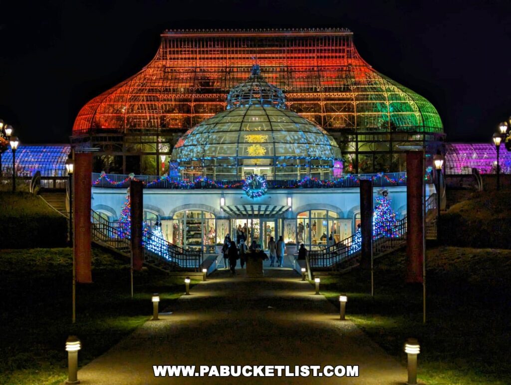 Nighttime view of the illuminated entrance to the Phipps Conservatory during the Holiday Magic Winter Flower Show and Light Garden in Pittsburgh, with the grand glasshouse structure lit in festive red, green, and blue lights, and visitors walking along the pathway adorned with Christmas decorations and sparkling trees.