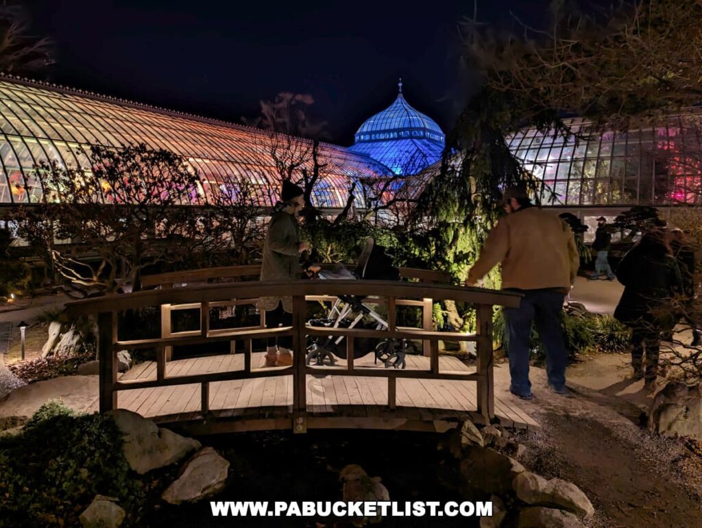 Visitors at night time standing on a traditional wooden bridge in the Japanese Garden section of the Phipps Conservatory, with the illuminated glasshouse and its colorful reflections in the background, creating a peaceful and picturesque scene during the Holiday Magic Winter Flower Show and Light Garden in Pittsburgh.