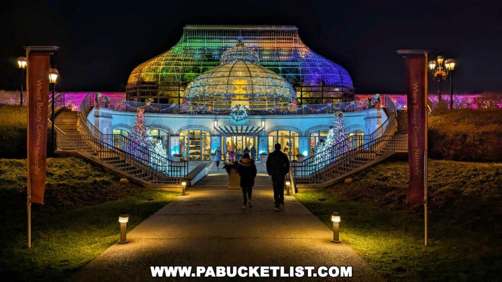 A couple walks towards the main entrance of the Phipps Conservatory at night, beautifully lit up with multicolored lights for the Holiday Magic Winter Flower Show and Light Garden in Pittsburgh, with the iconic glass dome structure radiantly illuminated in the background, showcasing the festive spirit and inviting ambiance.