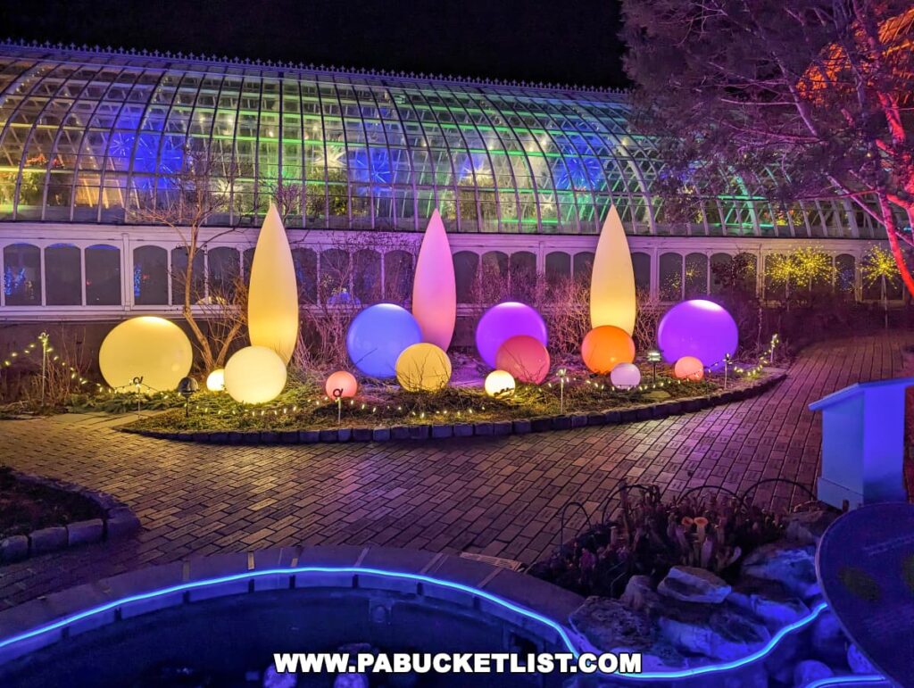 A serene outdoor scene at the Phipps Conservatory, featuring an array of oversized, illuminated bulbs in varying shades from warm yellow to cool purple, arranged in a row against the backdrop of the conservatory's glass greenhouse, which is lit from within, adding a magical glow to the night during the Holiday Magic Winter Flower Show and Light Garden.