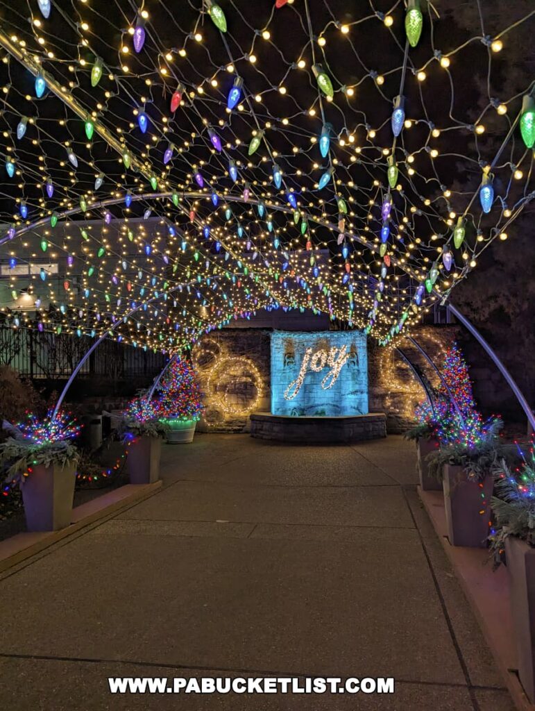 A festive walkway at the Phipps Conservatory Holiday Magic Winter Flower Show and Light Garden, adorned with a canopy of multicolored Christmas lights leading to a glowing 'Joy' sign set against a stone wall, creating a joyful and welcoming path for visitors during the holiday season in Pittsburgh.
