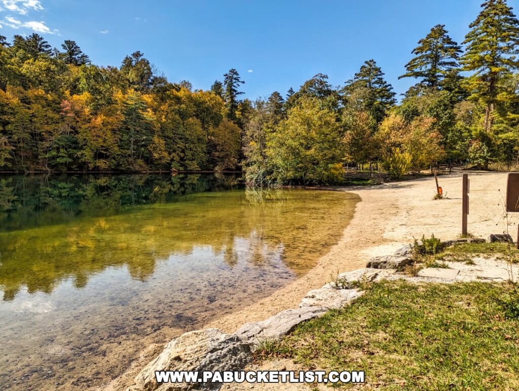 A peaceful beach at Fuller Lake in Pine Grove Furnace State Park, Cumberland County, PA, with a crescent of sandy shore curving into the reflective waters. The lake is bordered by forest displaying early autumn colors in a mix of green and turning leaves. A bright blue sky with wispy clouds overlooks the scene.