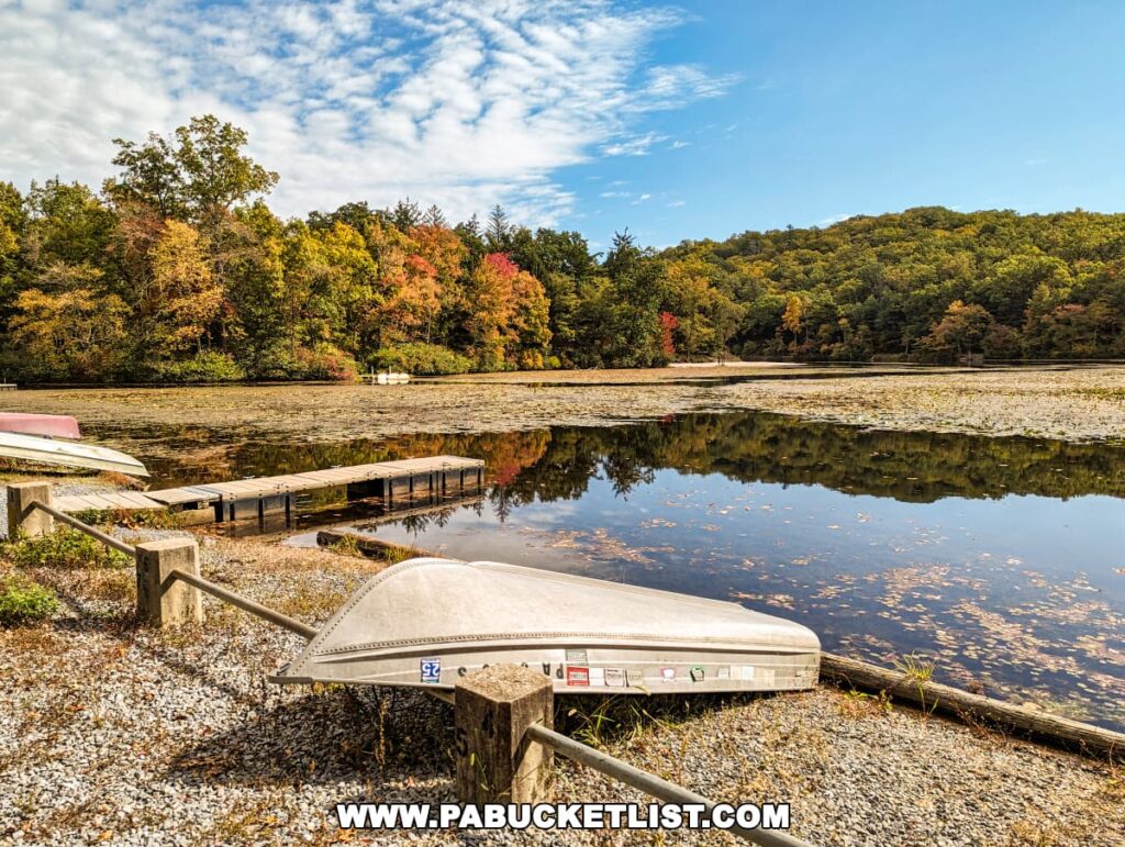 A lakeside view at Pine Grove Furnace State Park in Cumberland County, PA, featuring upside-down canoes on the gravel shore near a wooden dock. The reflective waters of Laurel Lake are partially covered with lily pads and fallen leaves, with a backdrop of forested hills displaying early signs of fall foliage. The sky is a mix of blue and cloud-streaked.