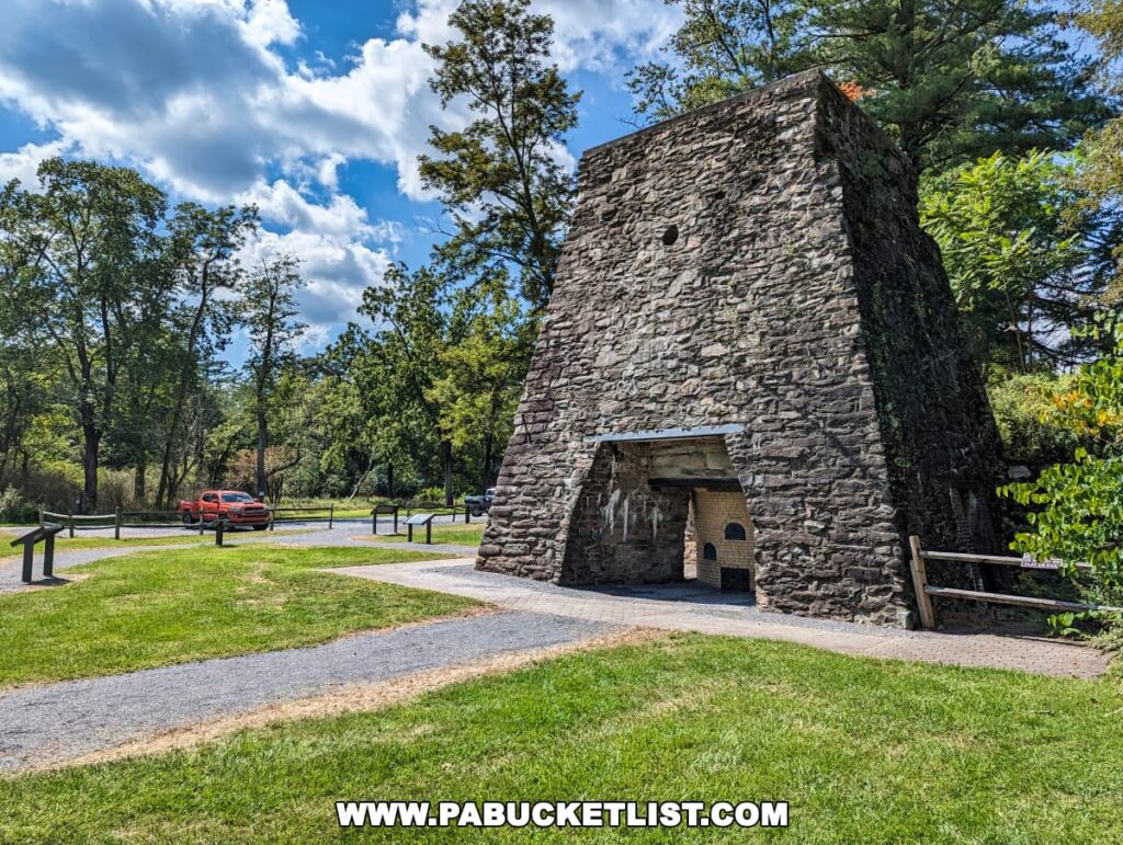 A historic stone furnace stack at Pine Grove Furnace State Park in Cumberland County, PA, stands under a blue sky with scattered clouds. The structure, made of large, irregular stones, features an archway at the base revealing a darkened interior. A wooden bench and walking paths are visible in the foreground, with lush green trees surrounding the area. A red car is parked in the distance, contrasting with the natural and historic setting.