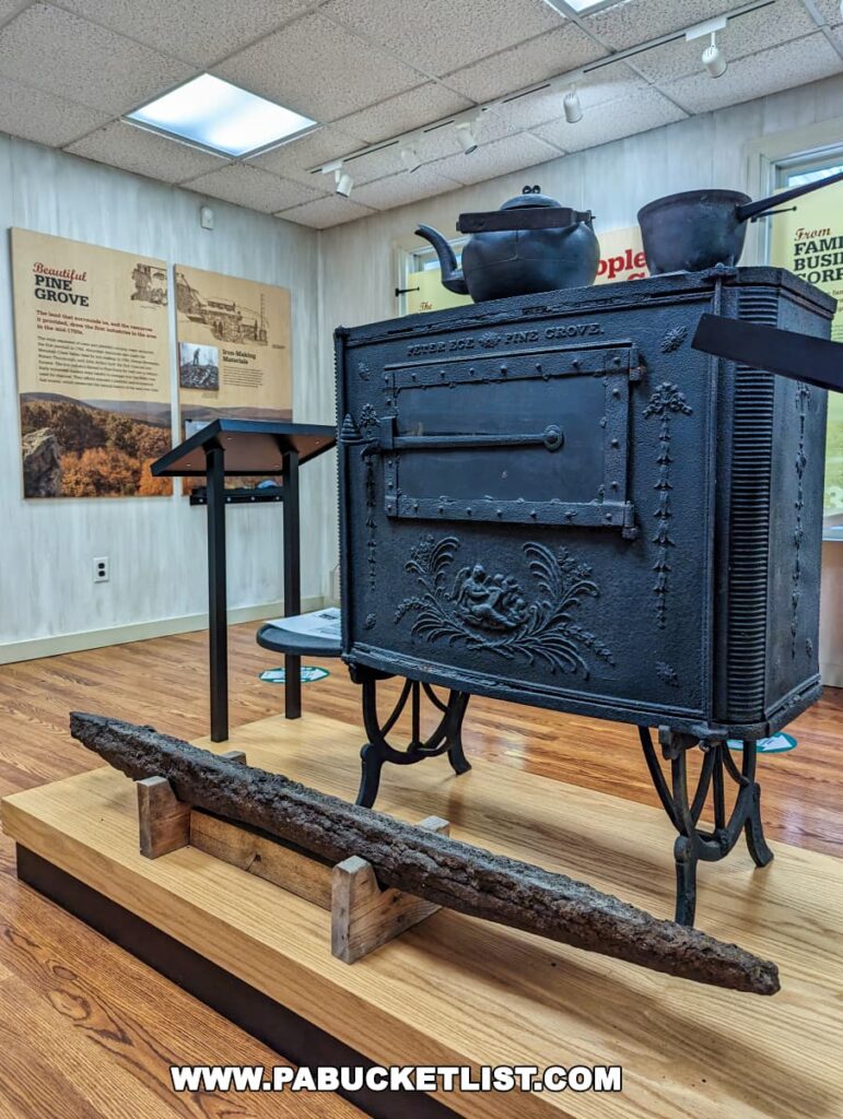 An antique iron stove from Pine Grove Furnace State Park in Cumberland County, PA, on display, with decorative detailing and a teapot and pan on top. In front of the stove is a long, rusted iron bar resting on wooden supports. The exhibit is in a room with informational posters on the walls, providing context about the park's history.