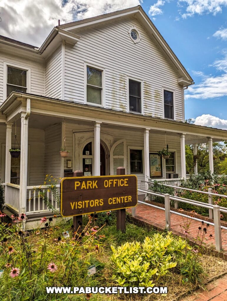 The Park Office and Visitor's Center at Pine Grove Furnace State Park in Cumberland County, PA, is a two-story white wooden building with a covered porch. A brown sign with yellow lettering directs visitors to the office. The building is set against a backdrop of partly cloudy skies, and the front garden is blooming with pink flowers and lush greenery.