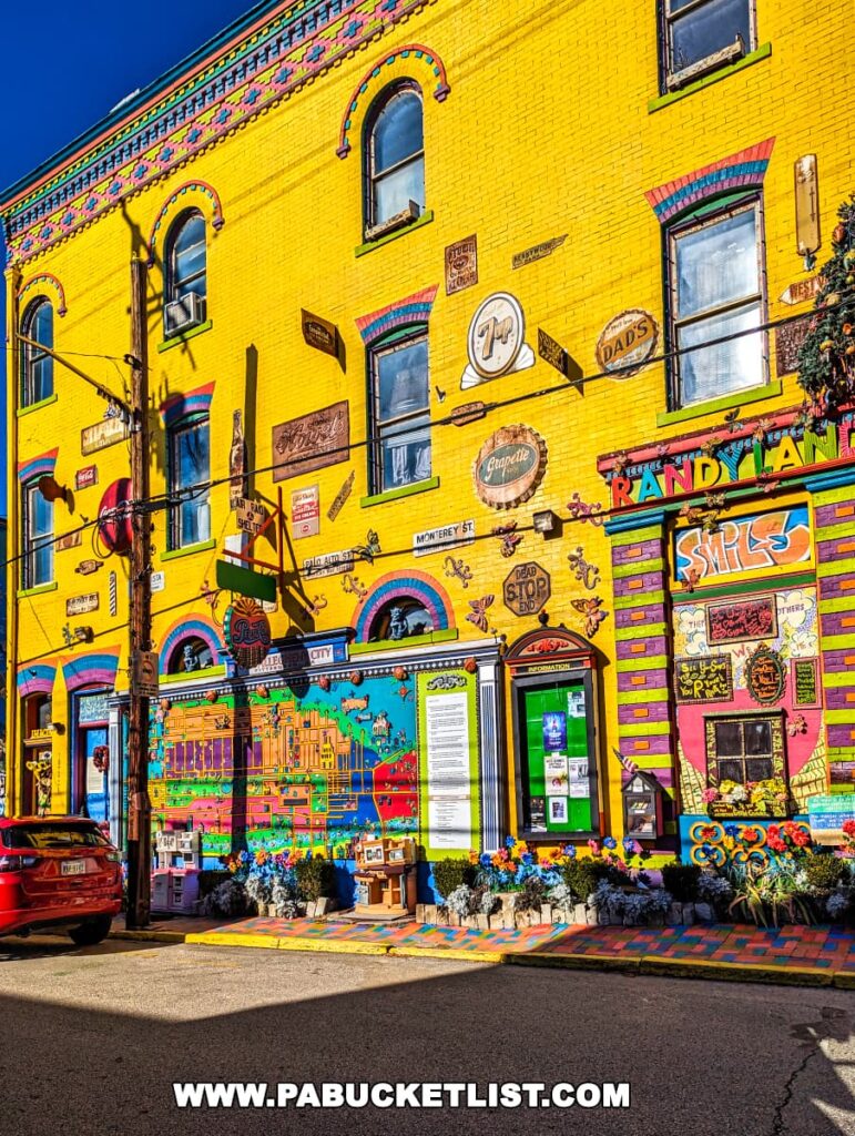 Street view of Randyland's vibrant facade in Pittsburgh with its iconic yellow walls adorned with colorful murals, vintage signs, and flower boxes under a clear blue sky.