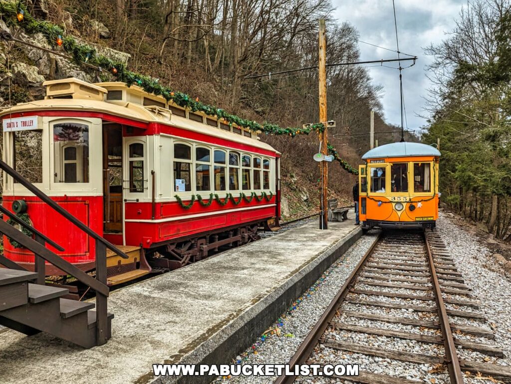 The scene shows two historical trolleys at the Rockhill Trolley Museum in Huntingdon County, PA. On the left, a red and white trolley, decorated with garlands and the words 'Santa's Trolley', is stationed at the platform. To the right, an orange trolley, numbered 355, sits on the tracks. Both trolleys are adorned with Christmas decorations, and a festive garland with lights stretches overhead between the trolleys against a cloudy sky backdrop.