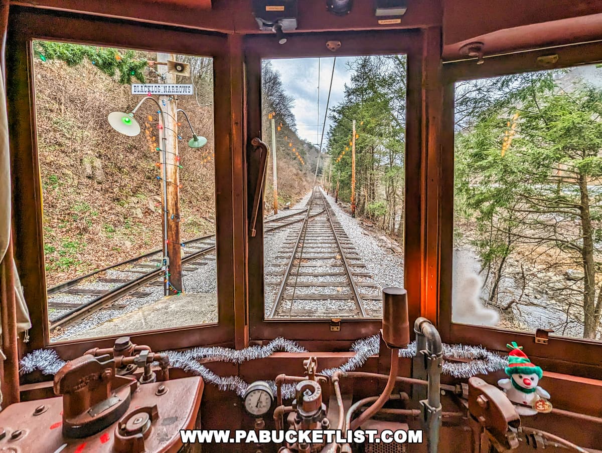 View from the operator's compartment inside a vintage trolley at the Rockhill Trolley Museum in Huntingdon County, PA, during the Santa's Trolley excursion. The controls of the trolley, including levers and gauges, are in the foreground, with a small festive snowman decoration. Through the front window, the trolley tracks extend straight ahead, flanked by bare winter trees and decorated with a string of colorful Christmas lights. A sign reading 'Blacklog Narrows' is visible to the left, adding to the historical charm of the scene.
