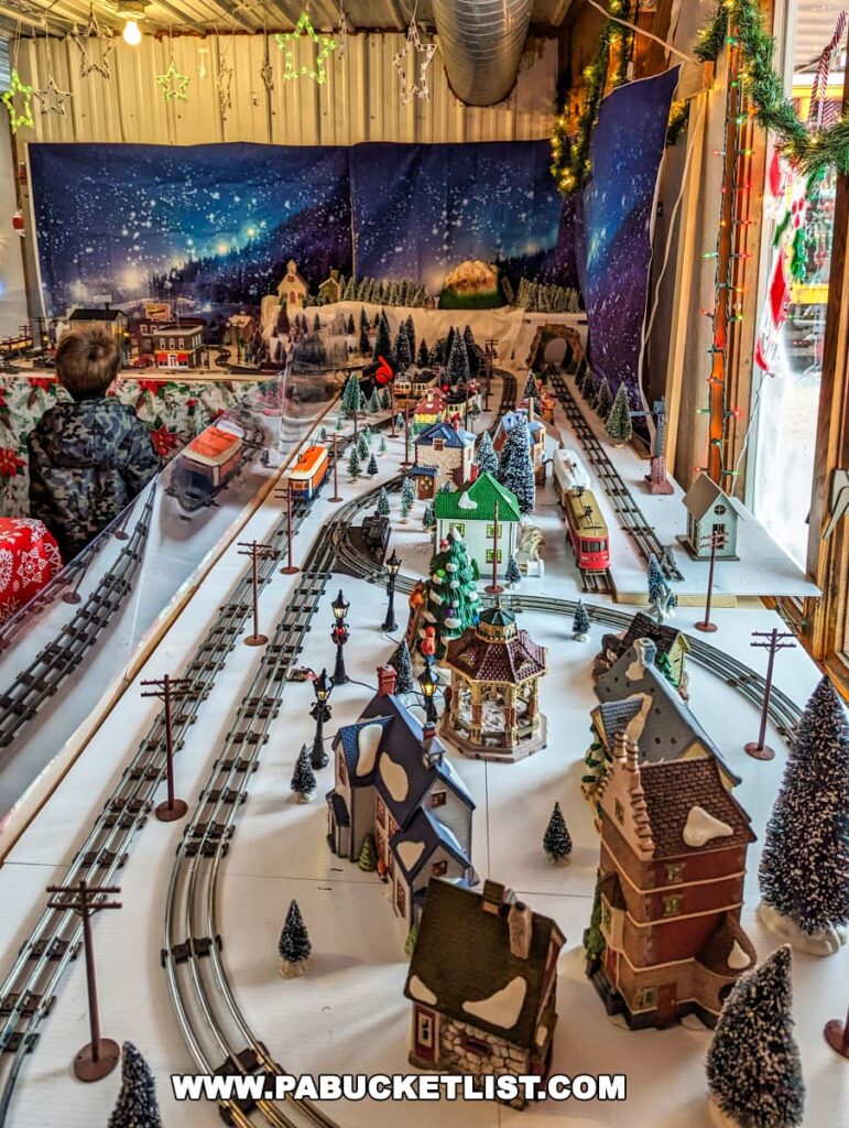 An elaborate miniature trolley display at the Rockhill Trolley Museum in Huntingdon County, PA, showing a snowy winter village scene. Multiple train tracks weave through the landscape, dotted with miniature houses, trees, and figurines. A backdrop painting of a night sky with stars and snowfall enhances the wintry effect. Festive decorations including garlands and lights are visible above the scene, and an onlooker is seen from the back, observing the moving trolleys.