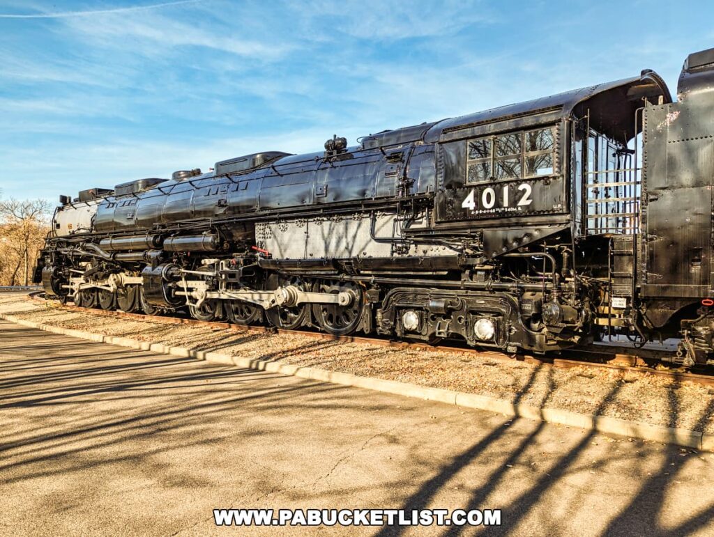 The Union Pacific "Big Boy" steam locomotive #4012 on display at the Steamtown National Historic Site in Scranton, Pennsylvania. This massive black locomotive with white numbering is a historic piece of American railroading, shown here with its intricate rods and wheels highlighted by the sunlight casting shadows on the ground. The locomotive sits on railroad tracks with bare trees and a blue sky in the background.