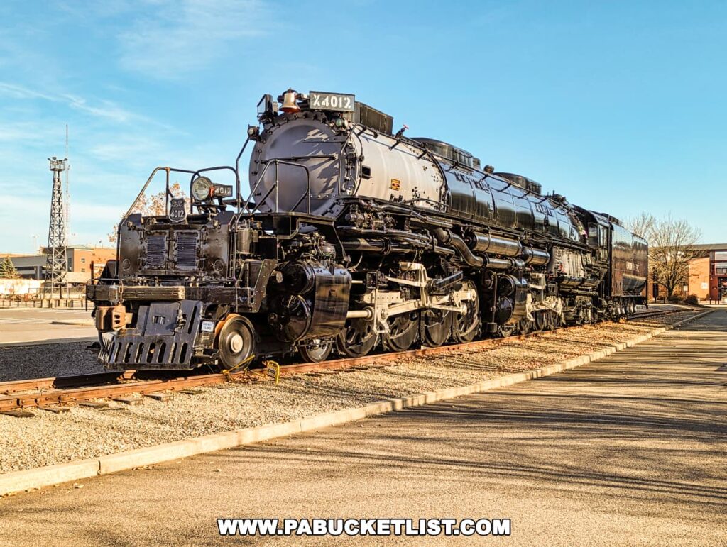 The iconic Union Pacific "Big Boy" steam locomotive #4012 is exhibited outdoors at the Steamtown National Historic Site in Scranton, Pennsylvania. The large, black locomotive with white lettering stands on railroad tracks, with its intricate machinery and massive wheels highlighted in the daylight. The background features industrial buildings and a clear blue sky.
