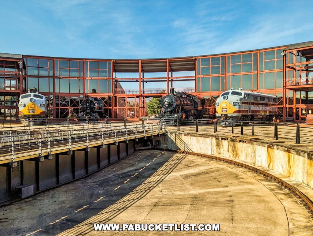 A large railway turntable at the Steamtown National Historic Site in Scranton, Pennsylvania, with two diesel locomotives, numbers 664 and 663, and a steam locomotive on the surrounding tracks. The turntable is in the foreground, showing its circular shape and the central pivot. The site features a modern architecture with a red and glass building in the background.