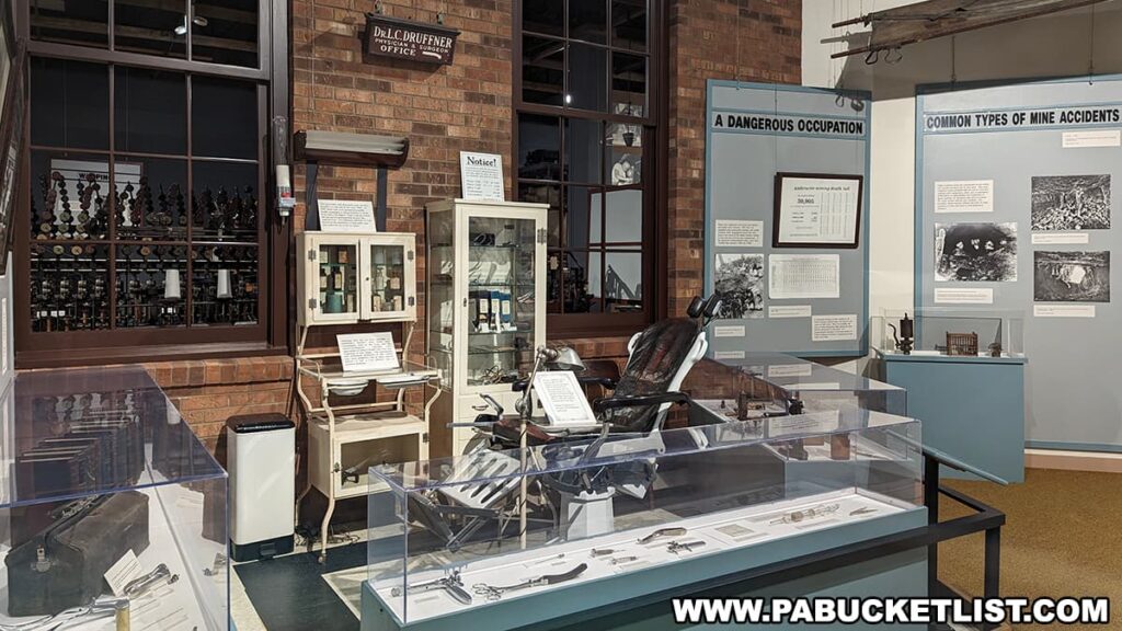 An exhibit at the Anthracite Heritage Museum in Scranton, PA, showcasing a recreated doctor's office from the coal mining era. The display includes a vintage medical cabinet, an old-fashioned dentist chair, and various medical instruments indicative of early 20th-century healthcare. Informational panels titled 'A Dangerous Occupation' and 'Common Types of Mine Accidents' provide historical context on the health risks associated with coal mining and the medical challenges faced by the miners.