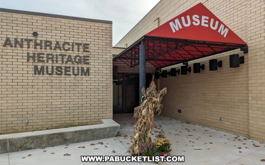 The entrance to the Anthracite Heritage Museum in Scranton, PA, featuring the museum's name in bold, raised lettering on a brick wall. A red awning with the word 'MUSEUM' welcomes visitors, and the entryway is adorned with a decorative cluster of dried cornstalks and autumnal flowers, indicating a seasonal display. The museum's facade gives a modern yet inviting impression to guests about to explore the history of coal mining in the region.