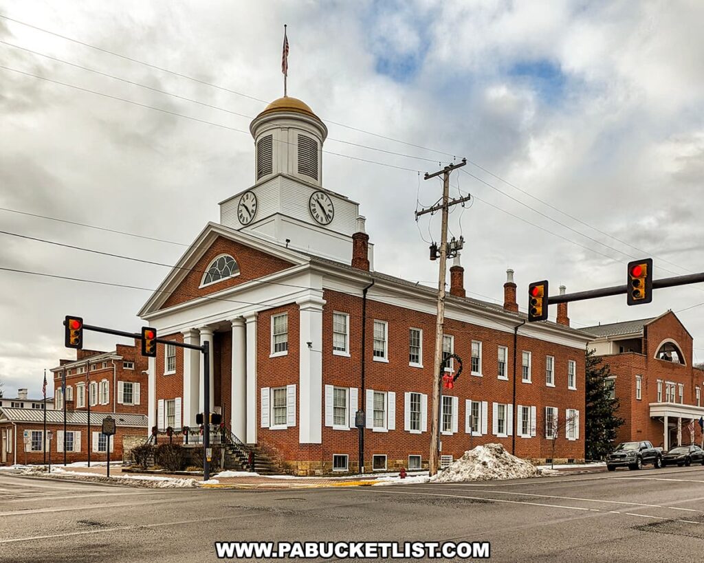 A daytime view of the Bedford County Courthouse in Pennsylvania, featuring its classic red brick facade and a prominent white columned entrance. The courthouse, topped with a white clock tower and a gold weathervane, sits at a street intersection with traffic lights showing red. Snow is piled on the street corners, and the sky is overcast, with the courthouse's American flag fluttering in the breeze.