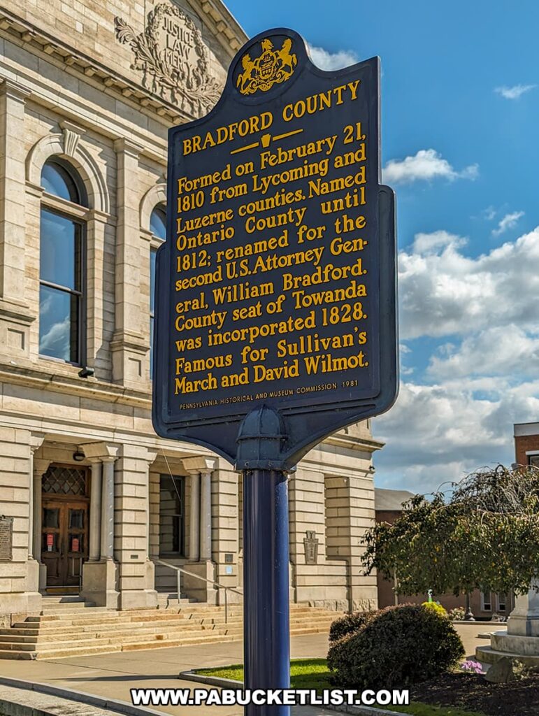 A historical marker in Bradford County, Pennsylvania, with the courthouse in the background. The blue and gold marker reads about the county's formation, name origin, and notable figures.